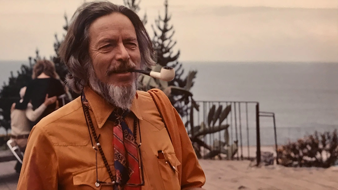Alan Watts in an orange shirt, holding a pipe beside the sea.