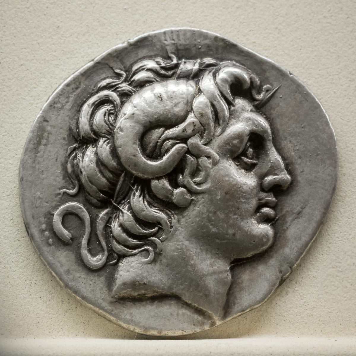 Silver coin showing Alexander’s head in profile facing right, with curling hair and a ram’s horn curving around his ear.