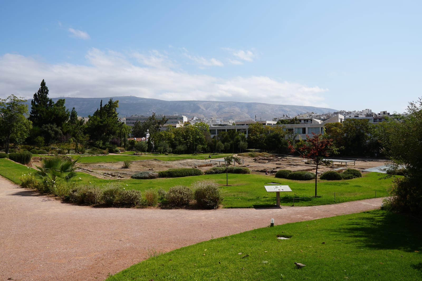 Excavated stone foundations in a grassy park in central Athens, with modern buildings and a long ridge of hills behind.