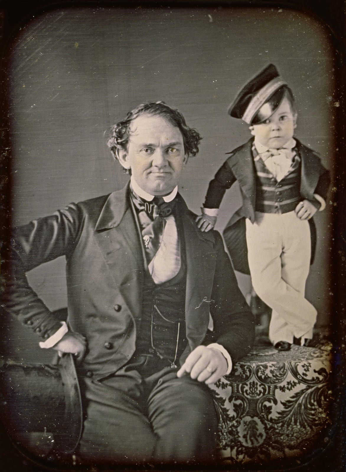 A daguerreotype of Barnum, seated, with a hand on his hip, and Charles Stratton standing on a draped table beside him in a cap, tailcoat, striped waistcoat and white trousers, one hand on Barnum’s shoulder.