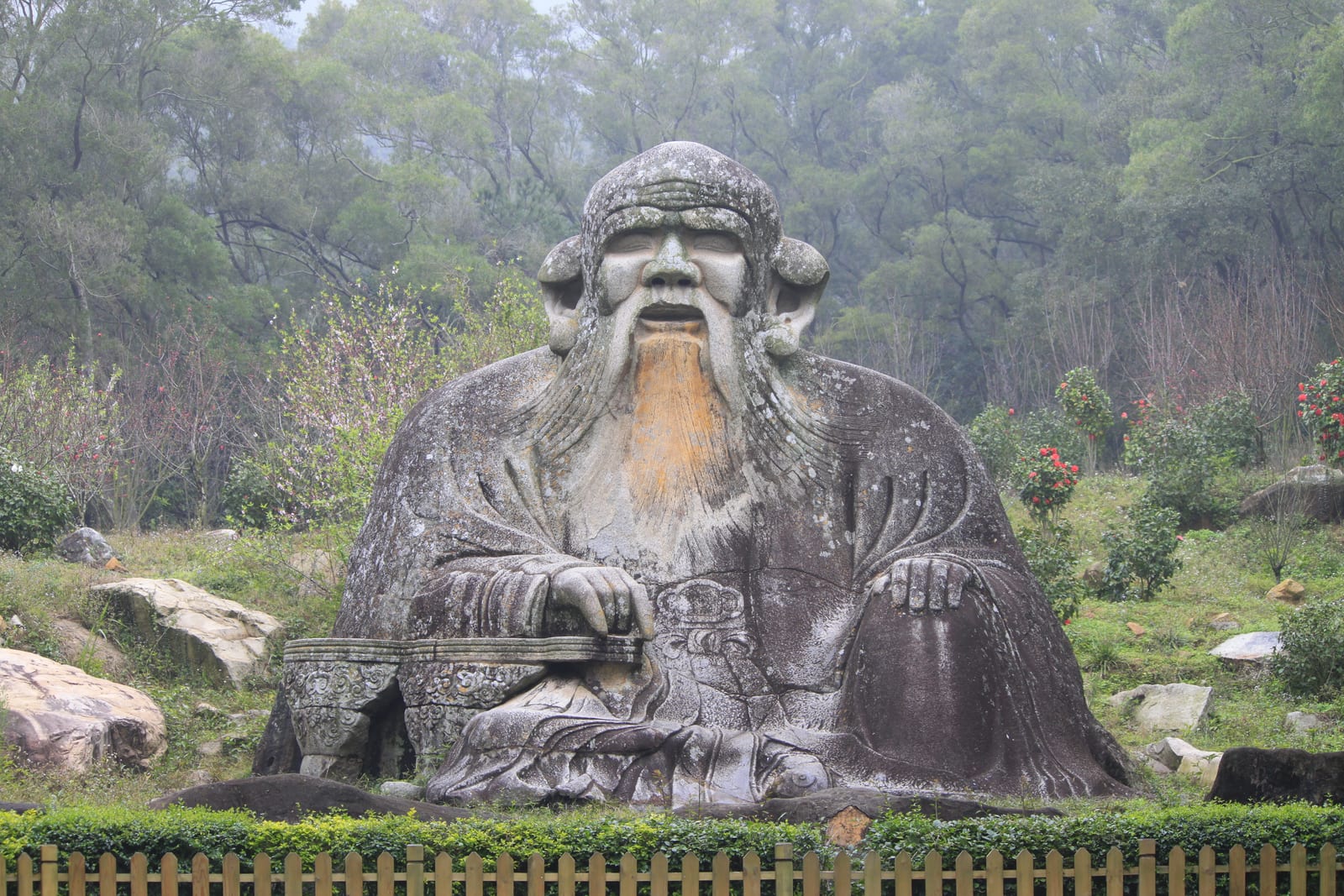 A huge weathered stone figure of a seated, long-bearded Laozi among trees on a misty hillside.