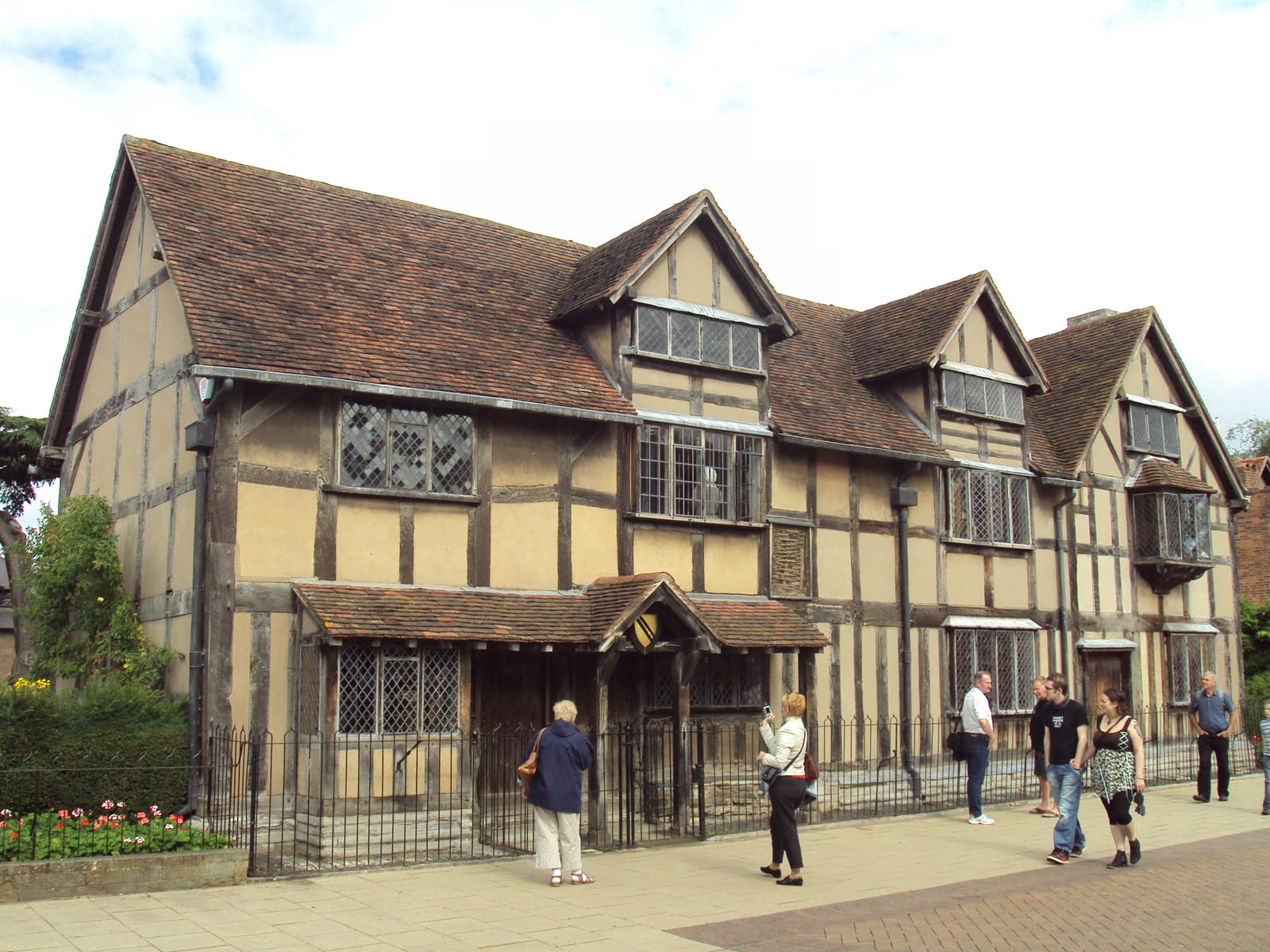 A long timber-framed house with gables and leaded windows on a pedestrian street, with visitors outside.
