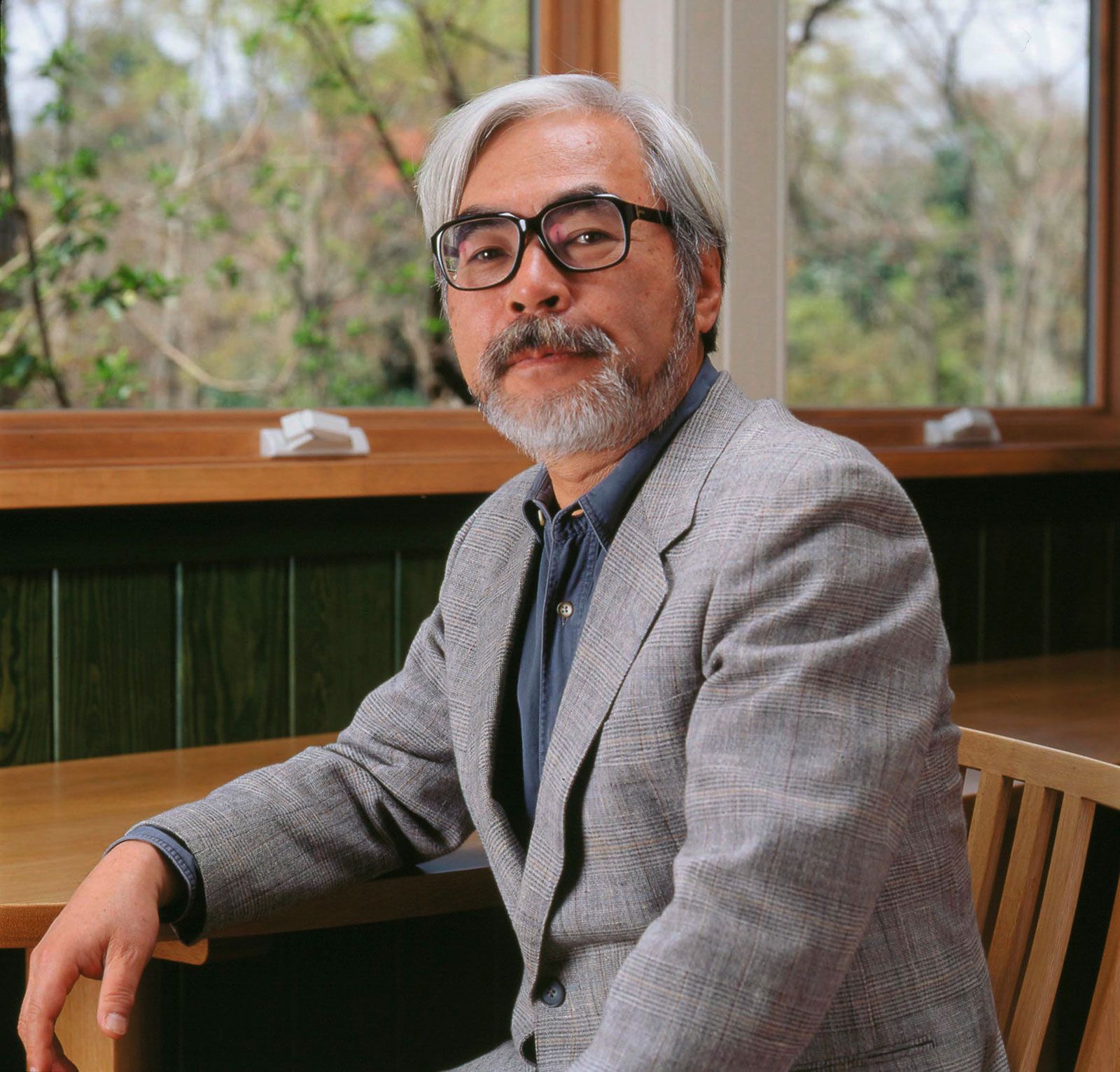 Hayao Miyazaki seated beside a window, wearing black glasses and a grey jacket.